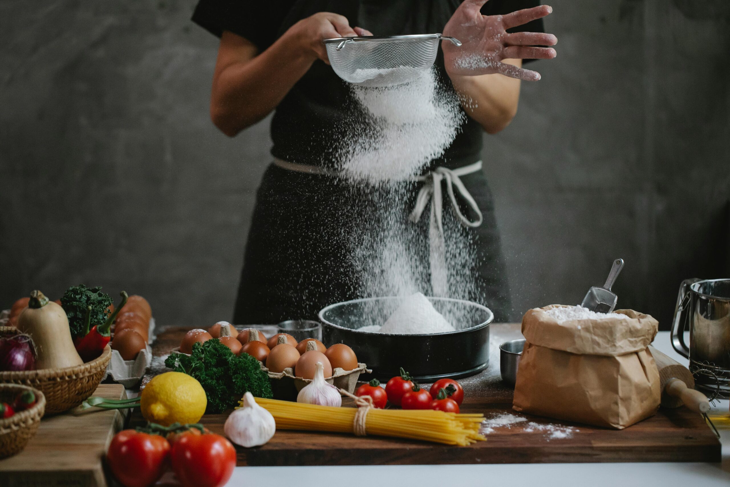 Person sifting flour over baking pan surrounded by eggs, vegetables, and kitchen ingredients on a rustic table


