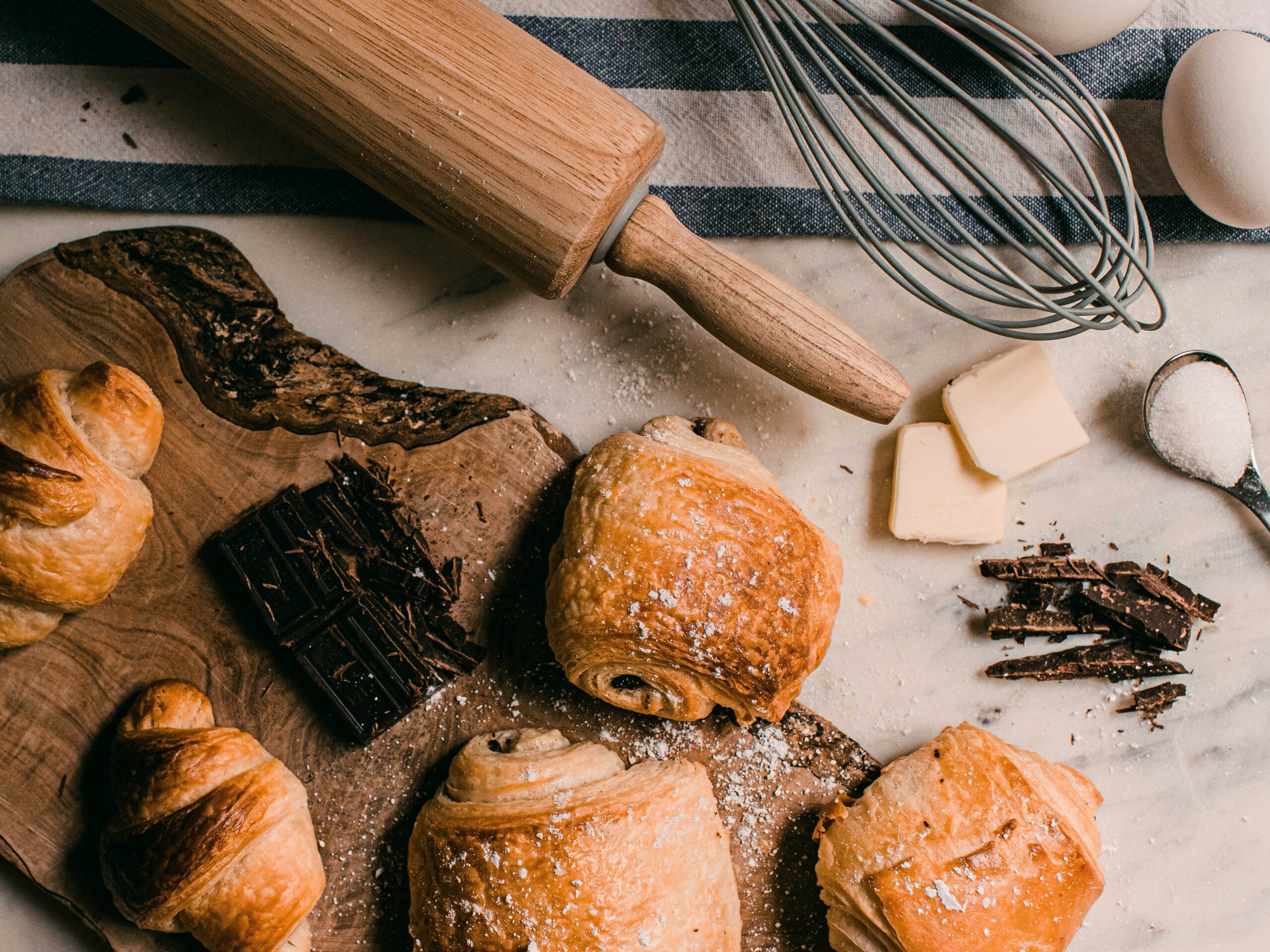 Fresh croissants and chocolate on a wooden board with rolling pin, whisk, butter, and baking ingredients on a marble surface